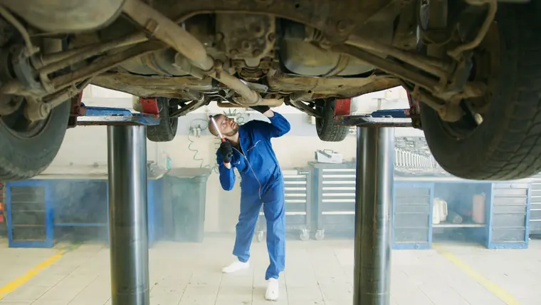 A vehicle raised on a two-post lift in a workshop while a technician inspects the underside